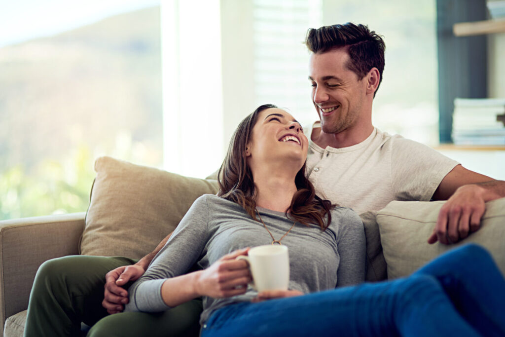Couple, drinking coffee and laugh in home living room, talking or bonding together to relax. Tea cup, happy man and woman in funny conversation on sofa for healthy relationship, connection or support